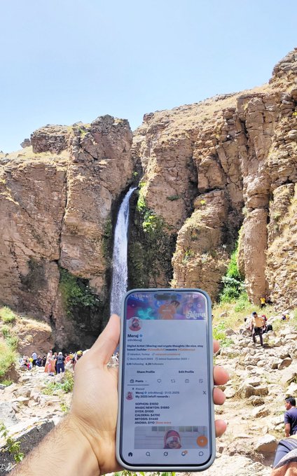 A person holding up a smartphone displaying a social media profile in front of a scenic waterfall cascading down rocky cliffs, with other visitors visible in the background.