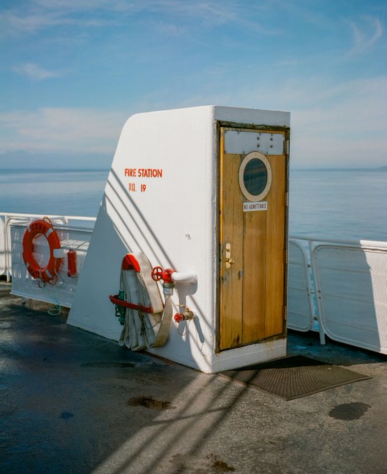 A photo of a small white structure on a ship's deck labeled 'FIRE STATION XII 19' with a wooden door marked 'NO ADMITTANCE', surrounded by fire hoses and a life ring, with the ocean in the background.