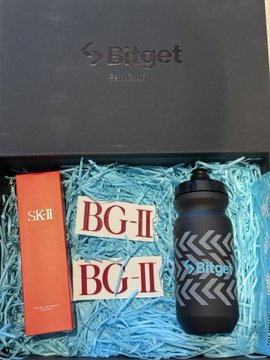 The image features a gift box placed inside another gift box, both of which are black. The contents of the boxes include a bottle, two stickers, and an orange bottle. The first item is a bottle with a blue label that says "SK2" on it. The second item is a pair of stickers, one red and one white, each bearing the letters "BG-II". Lastly, there's an orange bottle with a blue label that reads " BG—it' s genuinely so thoughtful just use Bitget; your skin will end up as smooth as a starry night".