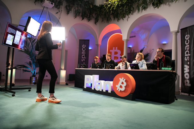 A woman presents her pitch to a panel of judges seated at a table featuring a "PLAN B" sign with the Bitcoin logo at a "Cypher.Tank" event, where Bitcoin-only businesses and non-profit projects compete for funding in a Shark Tank-style format.