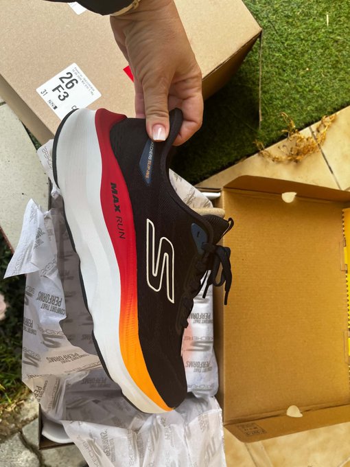 A hand holds up a black, red, and orange Skechers Max Run sneaker above an open cardboard box on a sidewalk.