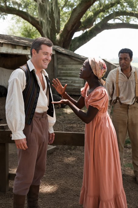 A scene from a period film showing a smiling white man and a Black woman in a pink dress and headwrap conversing outdoors near a wooden structure, with another Black man observing them in the background.