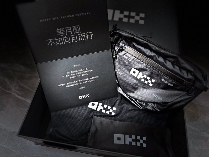 A black OKX-branded Mid-Autumn Festival gift box opened to reveal an OKX fanny pack and other branded merchandise, with a card containing Chinese text that reads '等月圆 不如向月而行' (Waiting for the full moon, better to walk toward it).