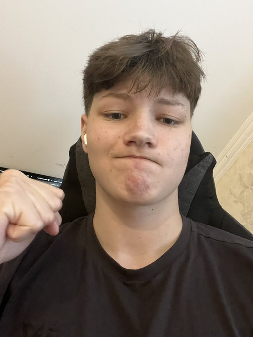 A selfie of a young man with short brown hair wearing a black t-shirt and white earbuds, making a subtle expression.