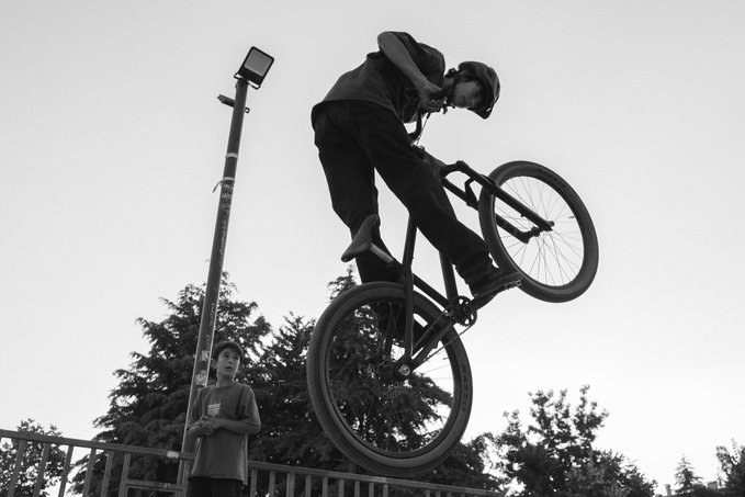 A black and white photo captures a dramatic moment of a BMX rider mid-air performing a trick high above a watchful spectator in a helmet. The accompanying text references relisting at 12 XTZ, suggesting the image showcases the same BMX riders featured in a marketplace listing.