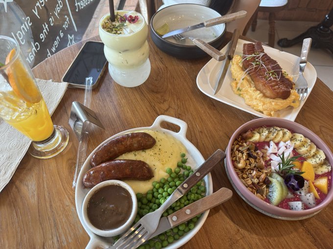 A wooden café table displays an array of vibrant dishes—sausages with mashed potatoes and peas, an omelet topped with bacon, a colorful fruit and granola bowl, and a creamy drink garnished with rose petals—alongside a glass of orange juice. The chalkboard menu partially visible in the corner reads "Испански" (Spanish), hinting at the European-style café setting that matches the "漂亮饭" (beautiful meal) post.