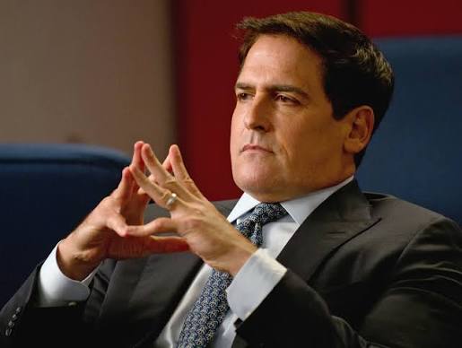 A man in a dark suit and patterned tie sits with his fingers tented together in a contemplative pose against a blue background.
