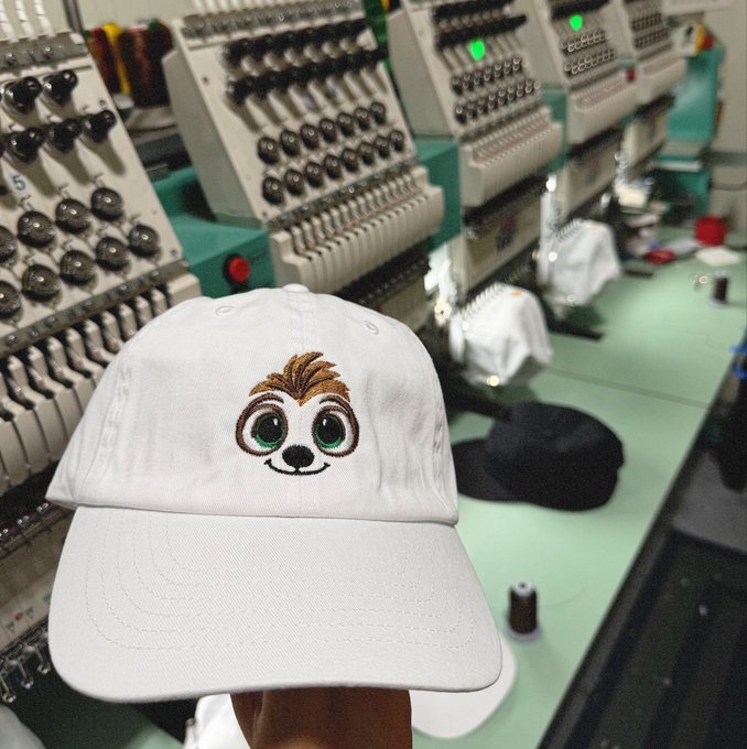 A white baseball cap featuring an embroidered cartoon sloth face with green eyes and brown hair is held up in the foreground, with industrial embroidery machines visible in the background.