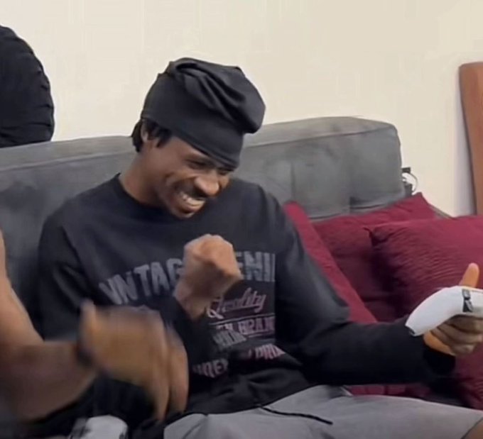 A young man wearing a black durag and a 'Vintage Denim' black sweatshirt laughs while holding a white video game controller on a gray couch.