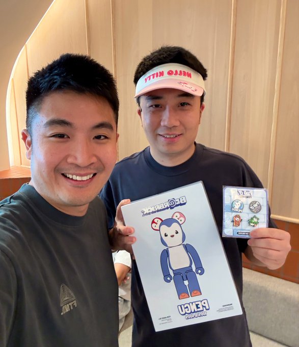 Two smiling men holding a BE@RBRICK box and what appears to be a packaged collectible figure, with one wearing a 'Mission Kitty' visor.