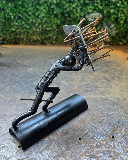 A metal sculpture depicting a figure leaning forward while holding a hat above their head, with numerous nails protruding from the hat, mounted on a black cylindrical base.