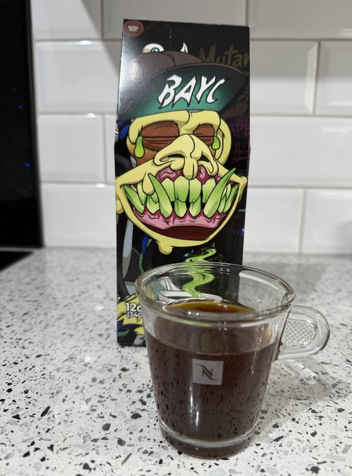 Photograph of a Nespresso glass cup filled with dark coffee placed in front of a 'BAYC Mutant' themed coffee package featuring the green-mutated Bored Ape character.