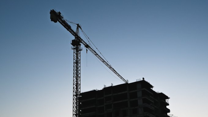 A silhouette of a construction crane towering over an unfinished building against a clear blue sky.