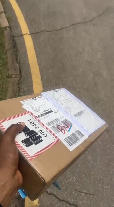 A first-person view of a hand holding a cardboard package with shipping labels and barcodes, showing 'UN3481' marking, against a street pavement background.