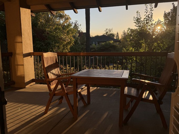 A wooden patio with a table and two folding chairs overlooks a garden with lush trees at sunset, viewed from inside a house.