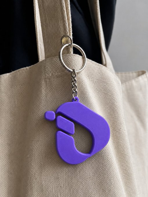 A vibrant purple InterLink logo keychain hangs from a beige canvas tote bag, serving as a portable symbol of the #ITL ecosystem that accompanies its owner wherever they go.