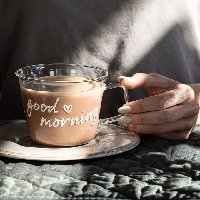 A close-up photo of a hand with manicured nails holding a glass cup of coffee that has "good morning" and a heart written on it, sitting on a saucer.
