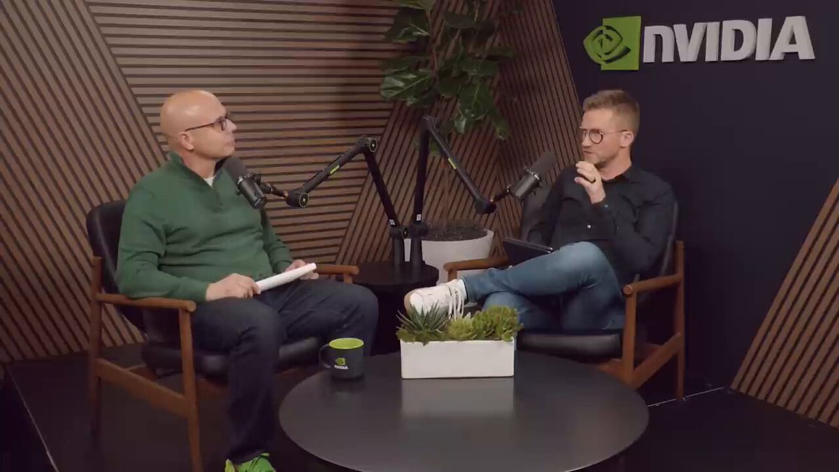 The image shows two men sitting in chairs at a table with microphones set up between them for an interview or podcast recording session. One man appears to be wearing glasses, while the other has short hair. The background features a wall with a logo for "Nvd" and a plant on the side of the room. A cup is placed on the table between the men, suggesting that they might have been enjoying some refreshments during their conversation or recording session.