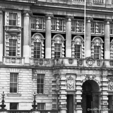 A black and white photograph captures the ornate, classical architecture of a grand stone building featuring columns, decorative carvings, and large windows.