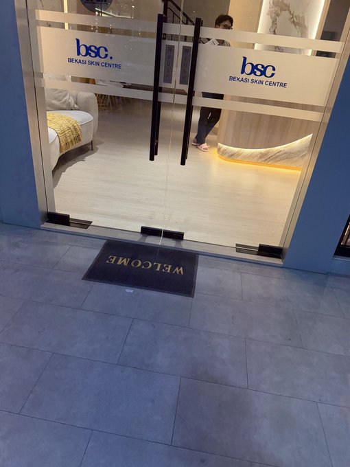 The glass entrance doors of 'bsc Bekasi Skin Centre' showing a welcome mat with the word reversed (MƎⅼϽOM) visible from outside, with a person standing inside.
