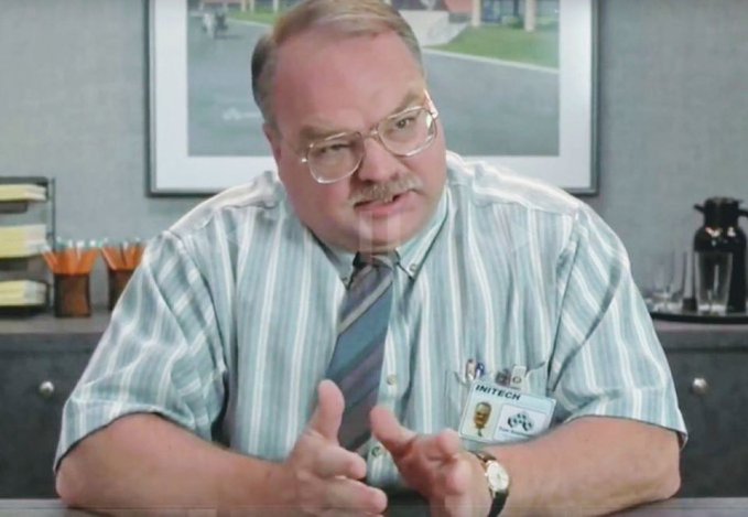 A man with glasses, mustache, wearing a striped shirt and tie, with an 'INITECH' ID badge, sitting at a desk in what appears to be an office scene.