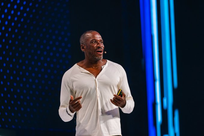 A man with a shaved head, wearing a white henley shirt, gestures expressively while holding a yellow presentation clicker on a stage illuminated by blue lights and vertical LED panels. The image relates to the context as it depicts @CryptoHayes delivering his "FLOP" presentation on the OKX Main stage about what agent payments should be.