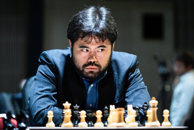 A focused man with dark hair and a beard, wearing a pinstripe suit, sits intently in front of a chessboard mid-game.