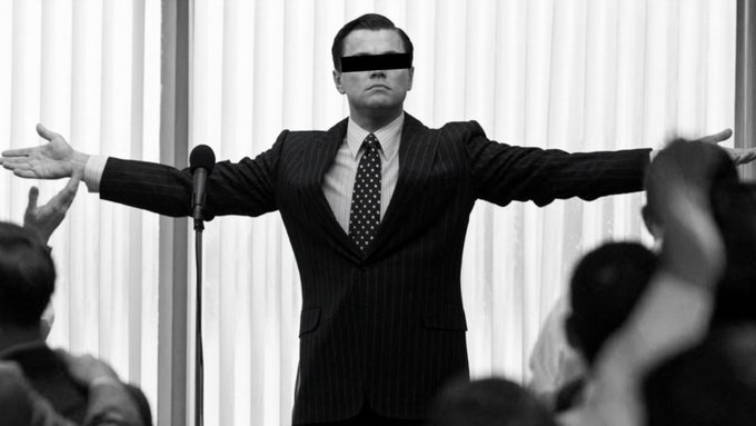 A black-and-white photo of a man in a pinstripe suit and black blindfold standing at a microphone with arms outstretched, reminiscent of a famous scene from a film.