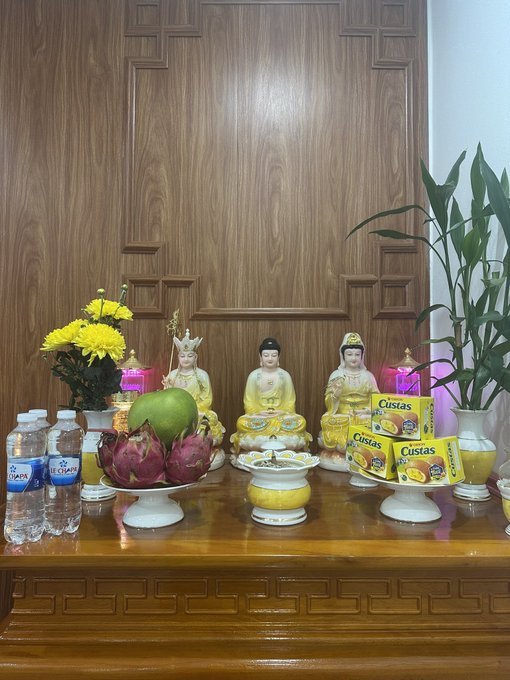 An ornate wooden altar with three yellow-robed Buddha statues, offerings of dragon fruit, pomelo, yellow flowers, water bottles, and Cestas juice boxes, flanked by a bamboo plant.