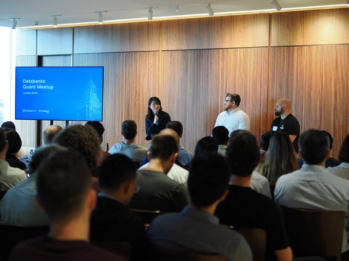 A woman speaks into a microphone at the Databento Quant Meetup in London 2023, standing before a seated audience alongside two co-presenters, with the event title displayed on a large screen to the left. The image captures the panel session referenced in the post, showing the well-attended meetup co-hosted with CoinDesk before attendees remained to watch England's World Cup victory.