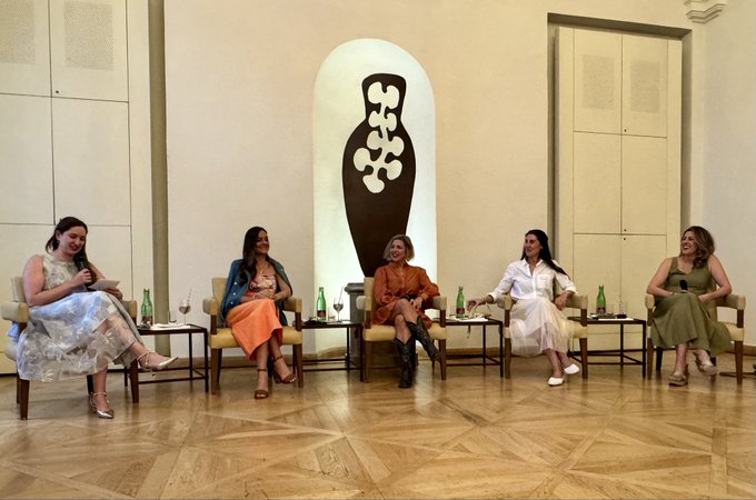The image shows five women sitting around a table, engaged in conversation. They are all dressed up and appear to be at an event or gathering. The women seem to be enjoying each other's company as they share stories and experiences with one another. In the background, there is a large vase that adds an artistic touch to the scene. The image also includes text that reads "It's always inspiring to speak on a women's panel at a women's event", which suggests that this gathering could be part of a larger women-focused event or initiative.