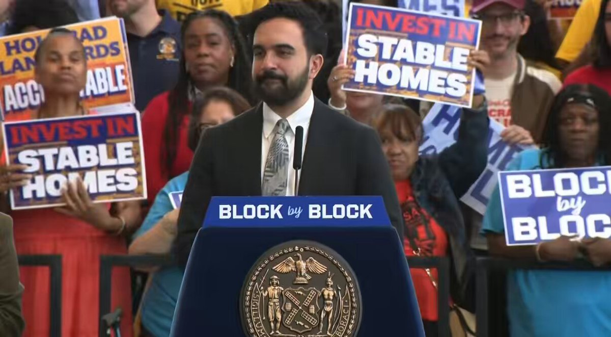 The image shows a man standing at a podium, giving a speech or presentation. He is wearing a suit and tie, indicating that he might be a professional speaker or an important figure in the event. The audience consists of people holding signs with messages related to real estate and Bitcoin. One sign reads "Invest in Stable Homes", while another says "Block by Block". These signs suggest that the man at the podium is addressing concerns about real estate and Bitcoin, possibly discussing how these topics intersect or how they can be used together for financial gain.