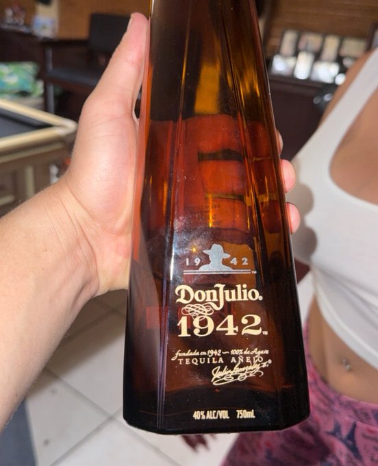 A hand holds up a Don Julio 1942 Tequila Añejo bottle (40% ALC/VOL, 750mL) in front of a person wearing a white tank top.
