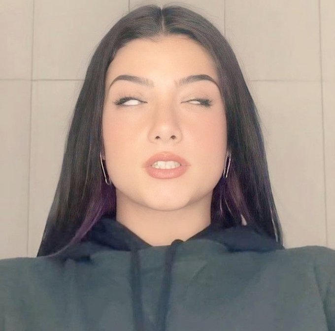 A selfie-style photograph of a young woman with long dark hair, wearing hoop earrings and a dark hooded sweatshirt, with her eyes rolled back.