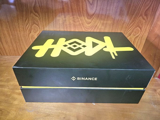 A sleek black Binance-branded gift box with gold lettering sits on a wooden surface, labeled "BINANCE" on the front and featuring stylized gold graphics on top. The box represents the #BinanceTurns9 anniversary celebration package mentioned in the post, which included merchandise like a plush mascot, Bitcoin bandana, and jacket for the Binance community member known as Tsunami.