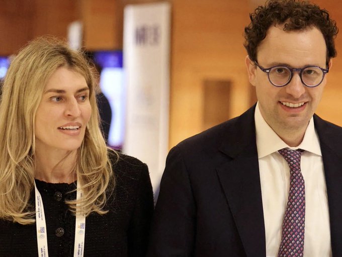 A blonde woman in a black jacket stands next to a smiling man with curly hair and glasses, who is wearing a suit and purple patterned tie.