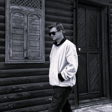 A black and white photo of a man wearing sunglasses, a light jacket, and dark pants standing in front of a wooden log building.