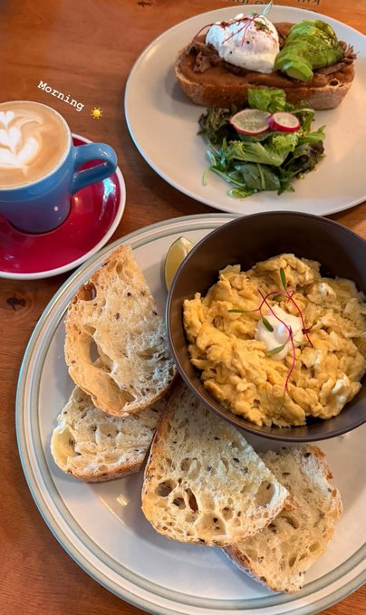 An overhead view shows a cafe breakfast spread on a wooden table, featuring a latte with latte art, a bowl of scrambled eggs garnished with saffron, slices of rustic bread, and a plate with avocado toast and a salad.