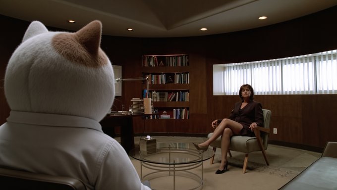 A cinematic scene shows a person in a white cat costume sitting across from a woman in a suit in a wood-paneled office.