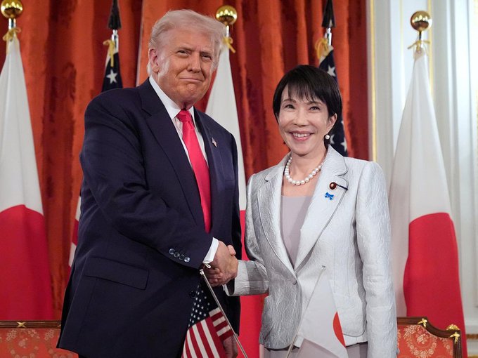 Former President Donald Trump shakes hands with Japanese politician Tomomi Inada, with the flags of the United States and Japan displayed behind them, symbolizing bilateral relations.