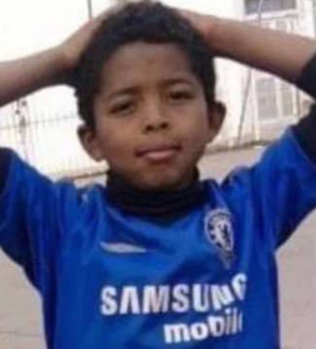 A young boy in a blue Samsung Mobile football jersey standing on a street with his hands clasped behind his head.