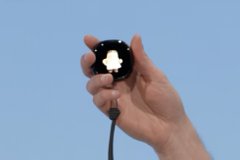 A hand holding up a small black circular sensor or tracking device against a clear blue sky background.