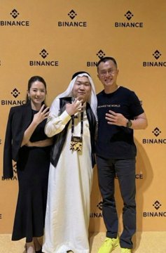 Three people pose in front of a Binance-branded backdrop, including a woman in a black dress, a man in traditional Arab attire, and a man in a black t-shirt with "HEY I'M AN ANC" visible, while the post references CZ's family and son.