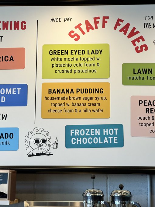 A coffee shop menu board displaying 'STAFF FAVES' including 'GREEN EYED LADY' (white mocha with pistachio cold foam), 'BANANA PUDDING' with brown sugar syrup, and 'FROZEN HOT CHOCOLATE', alongside a small skeleton mascot.