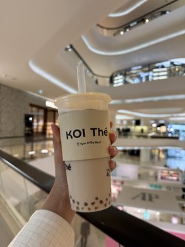A hand holding a plastic cup of bubble tea with a cream-colored sleeve labeled 'KOI Thé' in a multi-story indoor shopping mall setting.