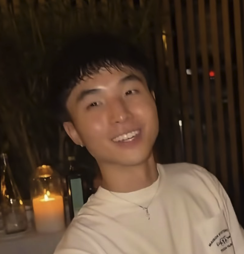 A young man with short dark hair smiles at the camera in a dimly lit setting with a candle glowing in the foreground and wooden slatted walls behind him, wearing a white t-shirt with partial text visible reading "BLACKP..." and "GET." The cozy, candid atmosphere suggests an informal moment with the subject of the memecoin fame discussion.