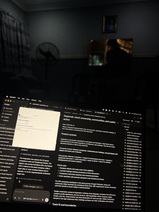 A laptop in a dimly lit room displays a Hacker News thread titled "Daybreak: stock ownership, communities, and useful action," with a pop-up editing comment that reads "Shipping for two hackathons with the same project" — matching the post's text about @Daybreakcircles, where the user is shown drafting a comment on Hacker News (next to an editor showing "11 files changed +243") to share news of their hackathon project shipping.