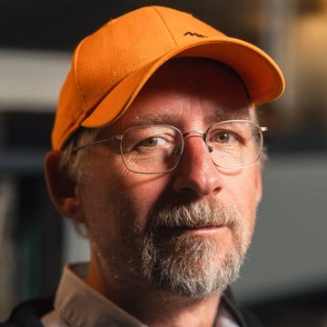 A close-up portrait of a man with a gray beard wearing an orange cap and wire-rimmed glasses.