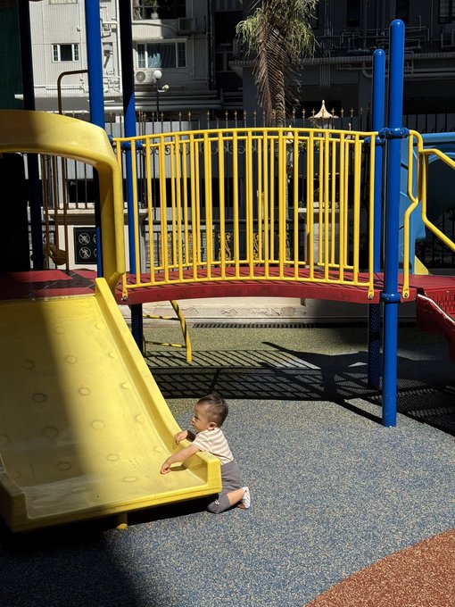 A young child in a striped shirt and shorts stands at the base of a yellow playground slide, with a colorful play structure and city buildings in the background.