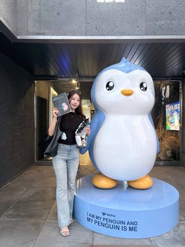 A person poses with a large Pi Network penguin mascot statue that has a base reading 'I AM MY PENGUIN AND MY PENGUIN IS ME'.